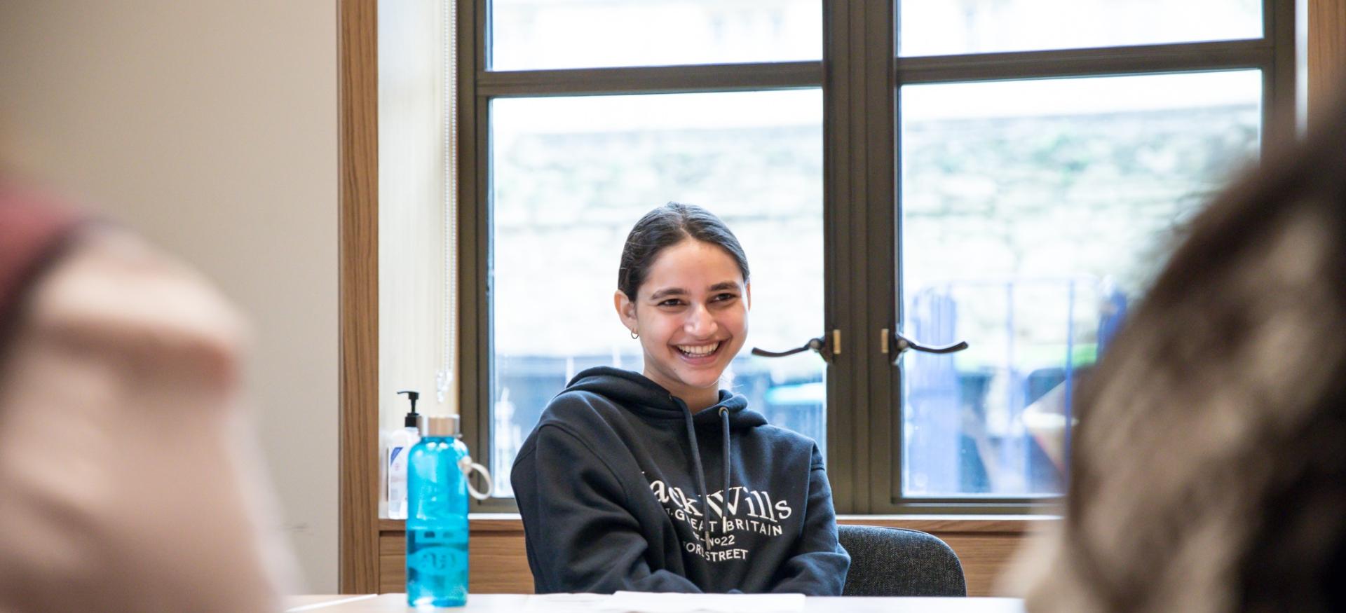 A student sits at a table during an academic discussion, smiling.