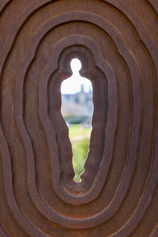 a detail of the spyhole in the gate designed by Antony Gormley. It is the outline of a person with hands at their sides. 