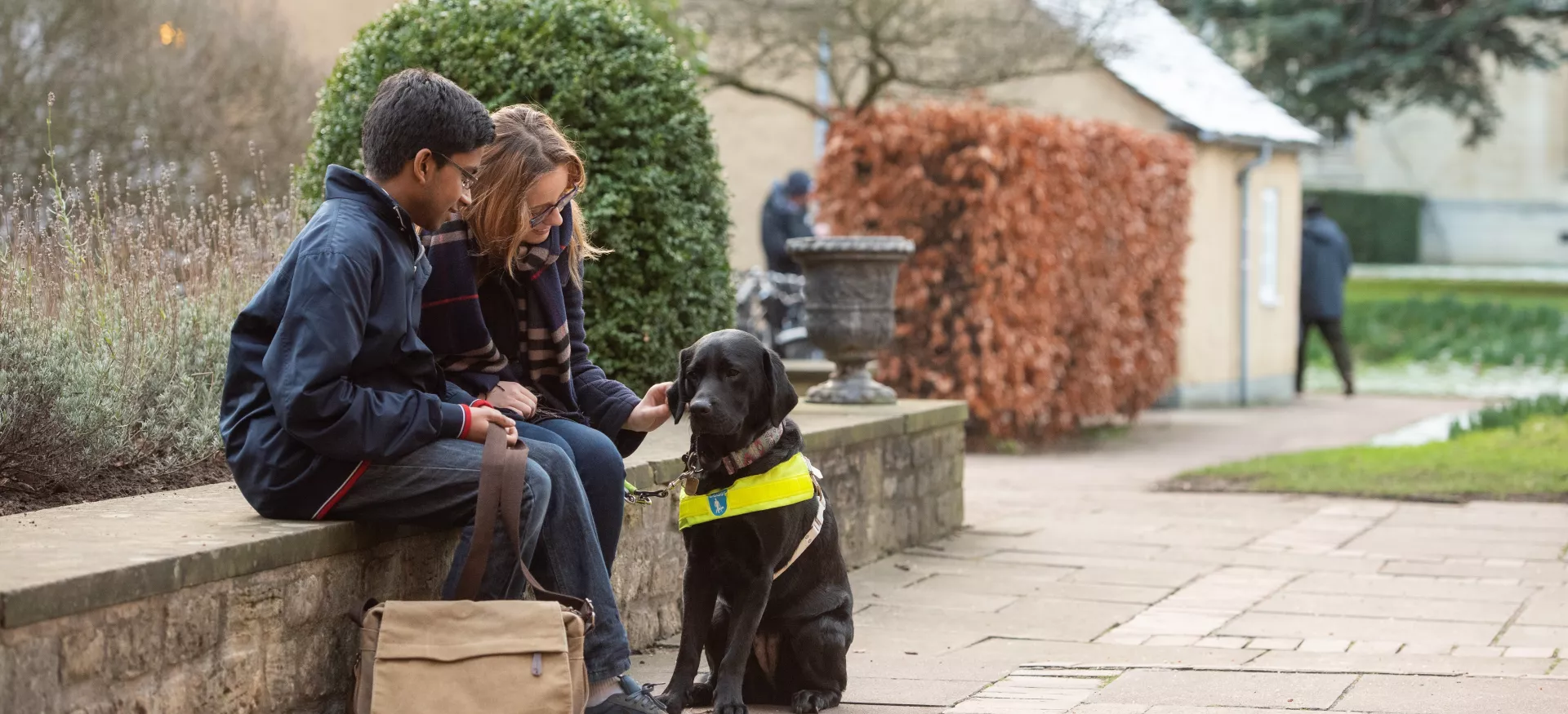 Two students, one with a guide dog, sit on a wall in Trinity College. 