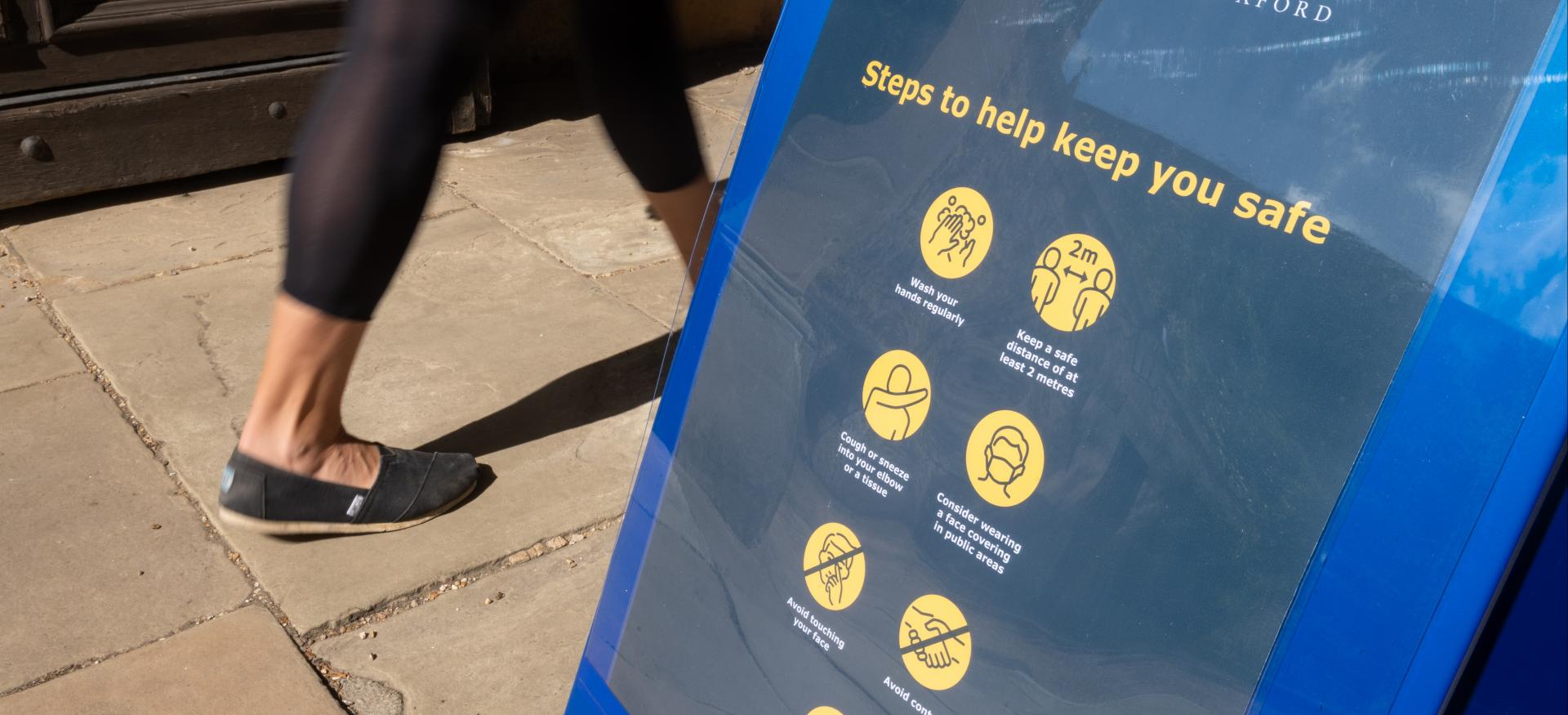 A woman's legs in motion walk past a sign saying 'Trinity College: steps to help keep you safe'