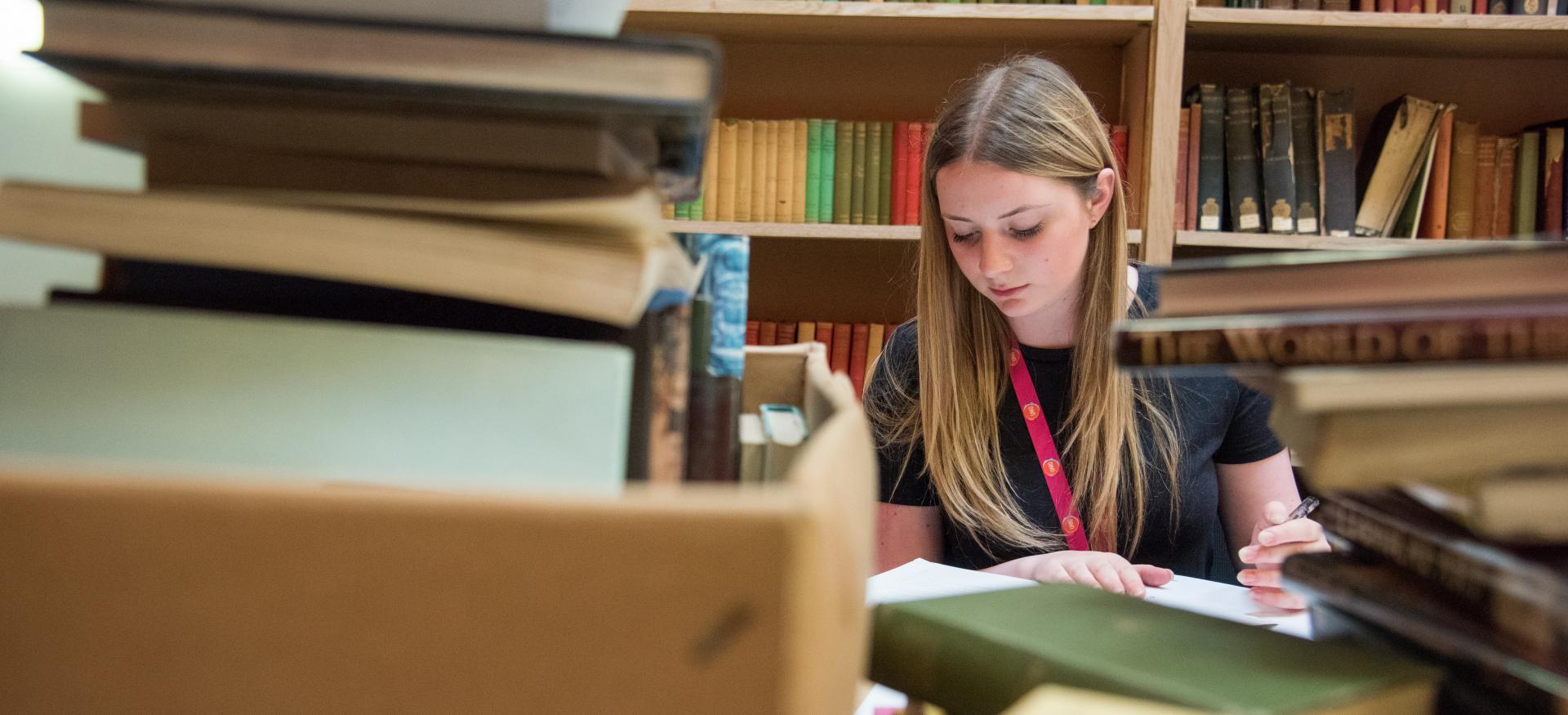 A student studies among a pile of books and folders with a bookshelf in the background.