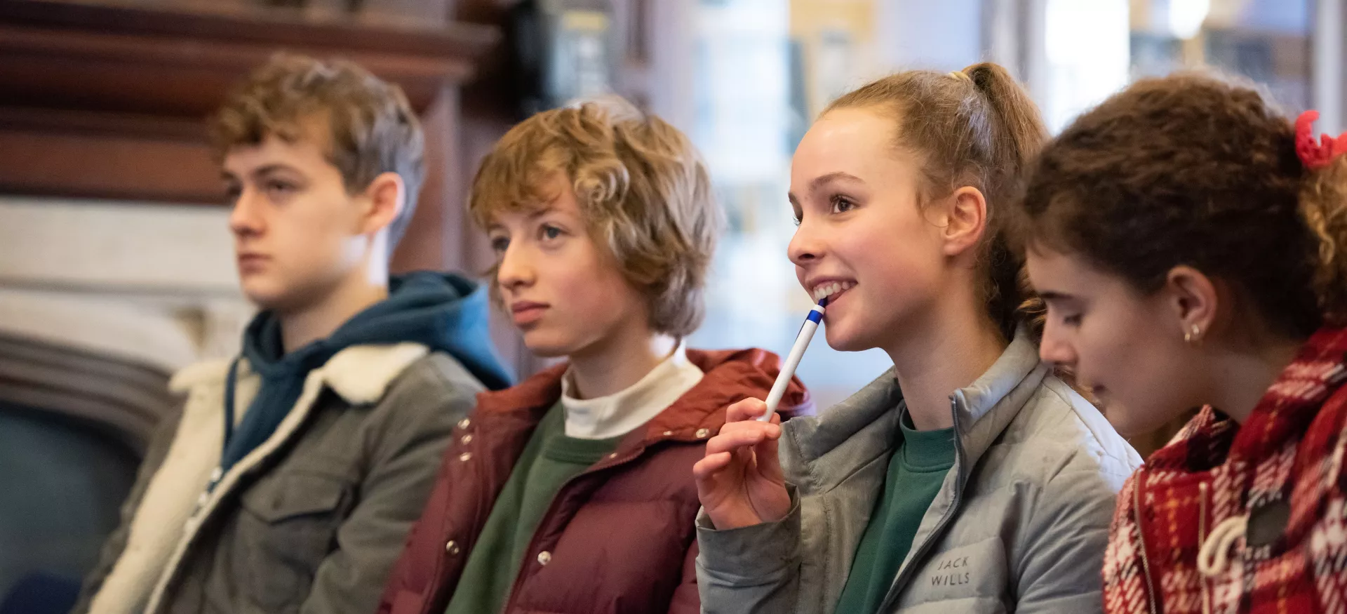 A group of secondary school students sit in a row listening to someone speak.