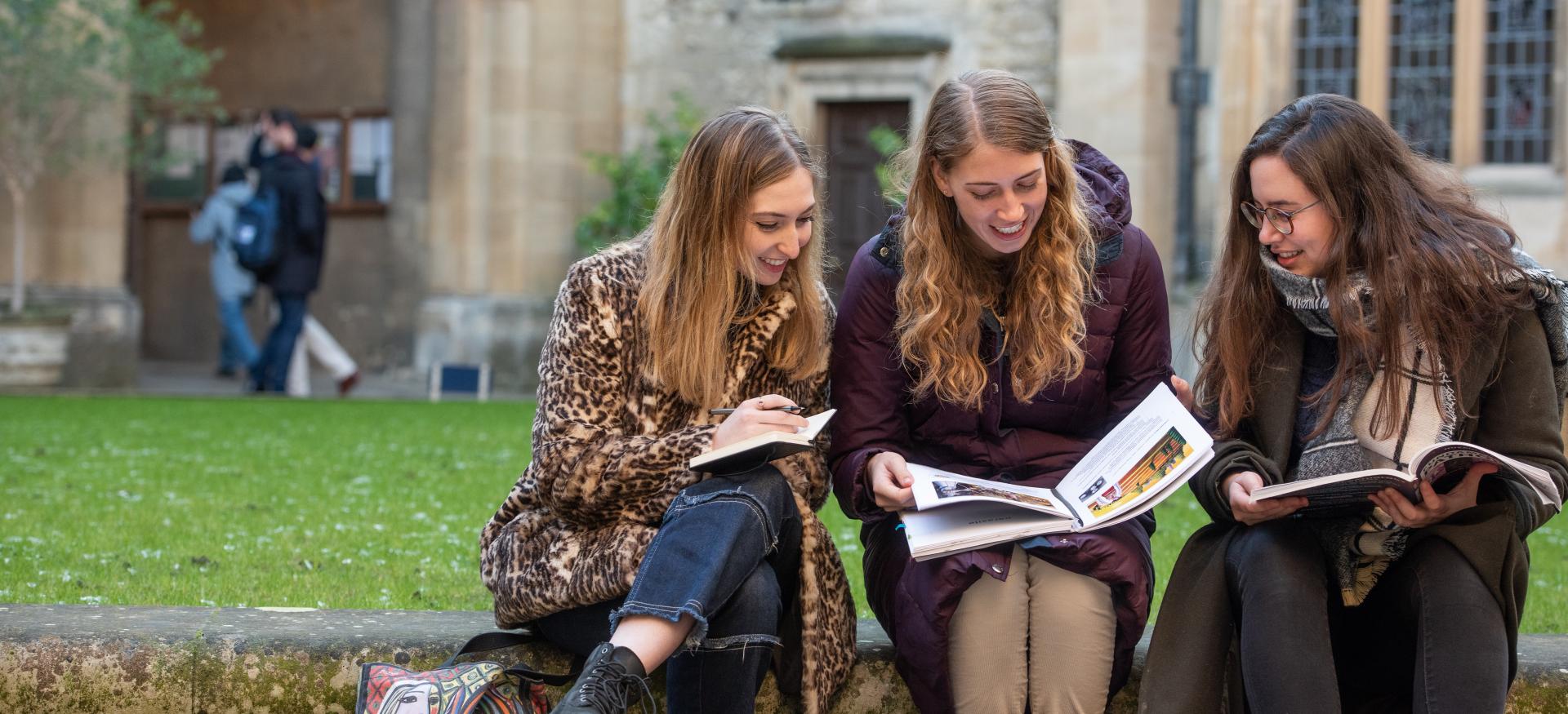 Three female Trinity college postgraduate students sit consulting books in one of the college quads.