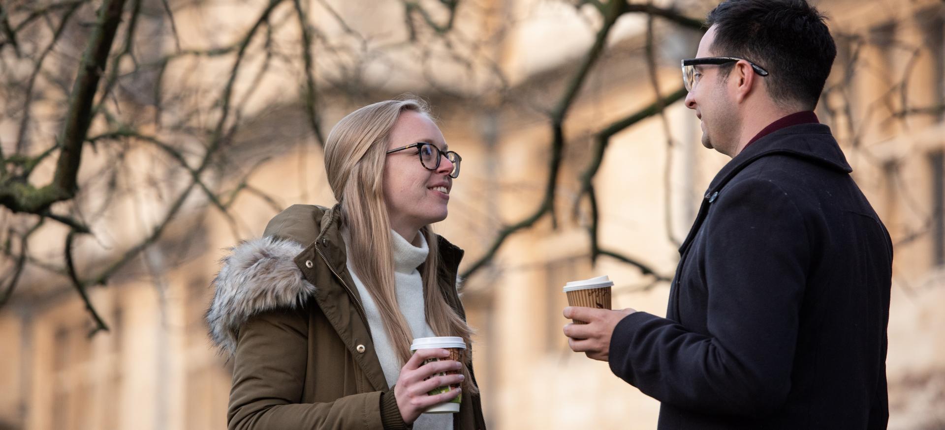 A pair of Trinity College postgraduates stand in profile holding coffee cups outside in the front quad.