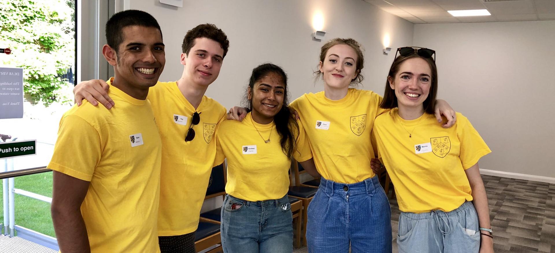 A group of five male and female Trinity students in yellow Open Days t-shirts stand with arms around each other at an Open Day.