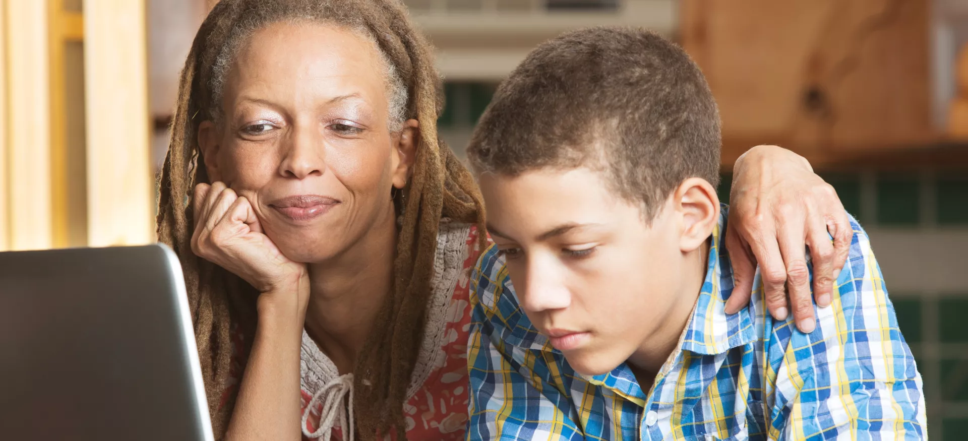 A mother overlooks as her teenaged son uses his laptop to study in their kitchen