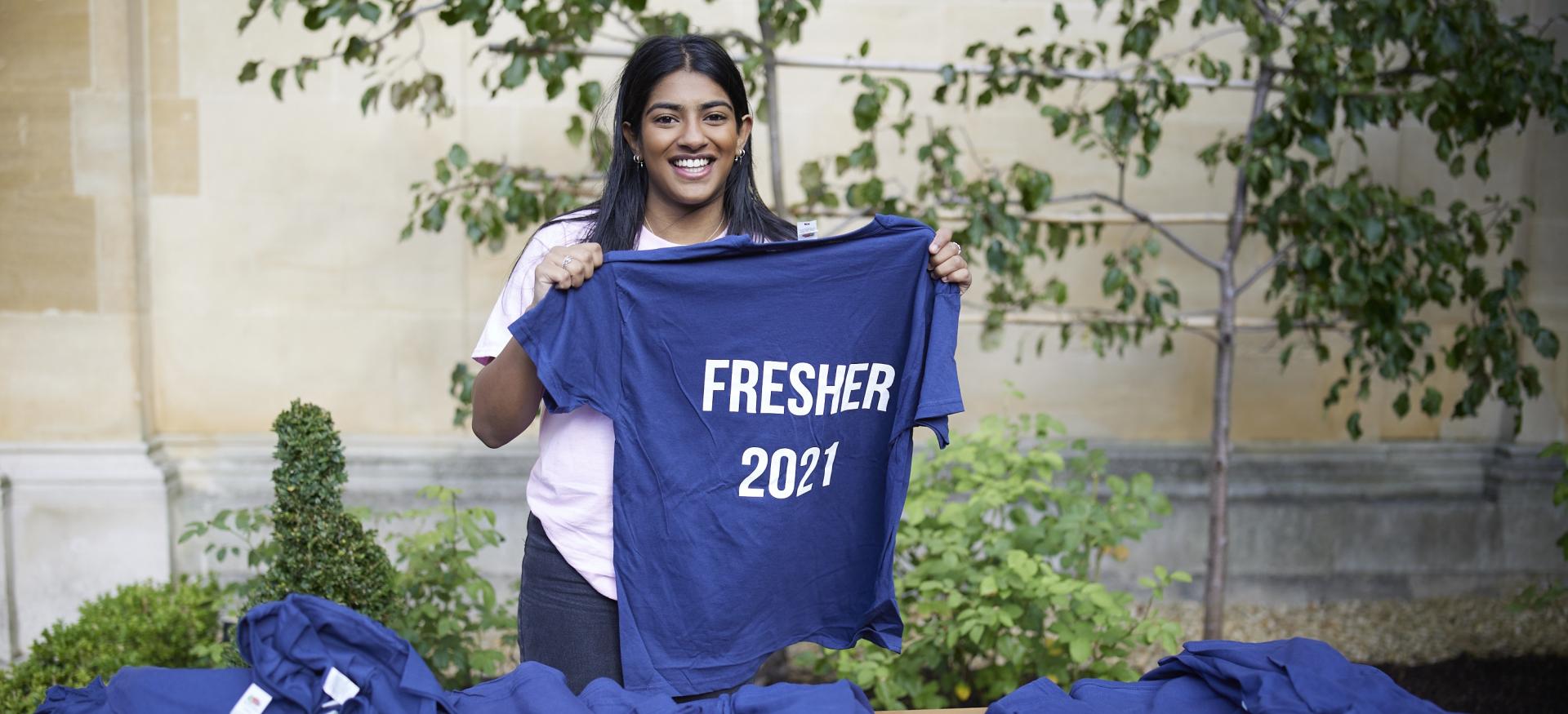 A female student stands holding a t-shirt that reads 'fresher 2021'.