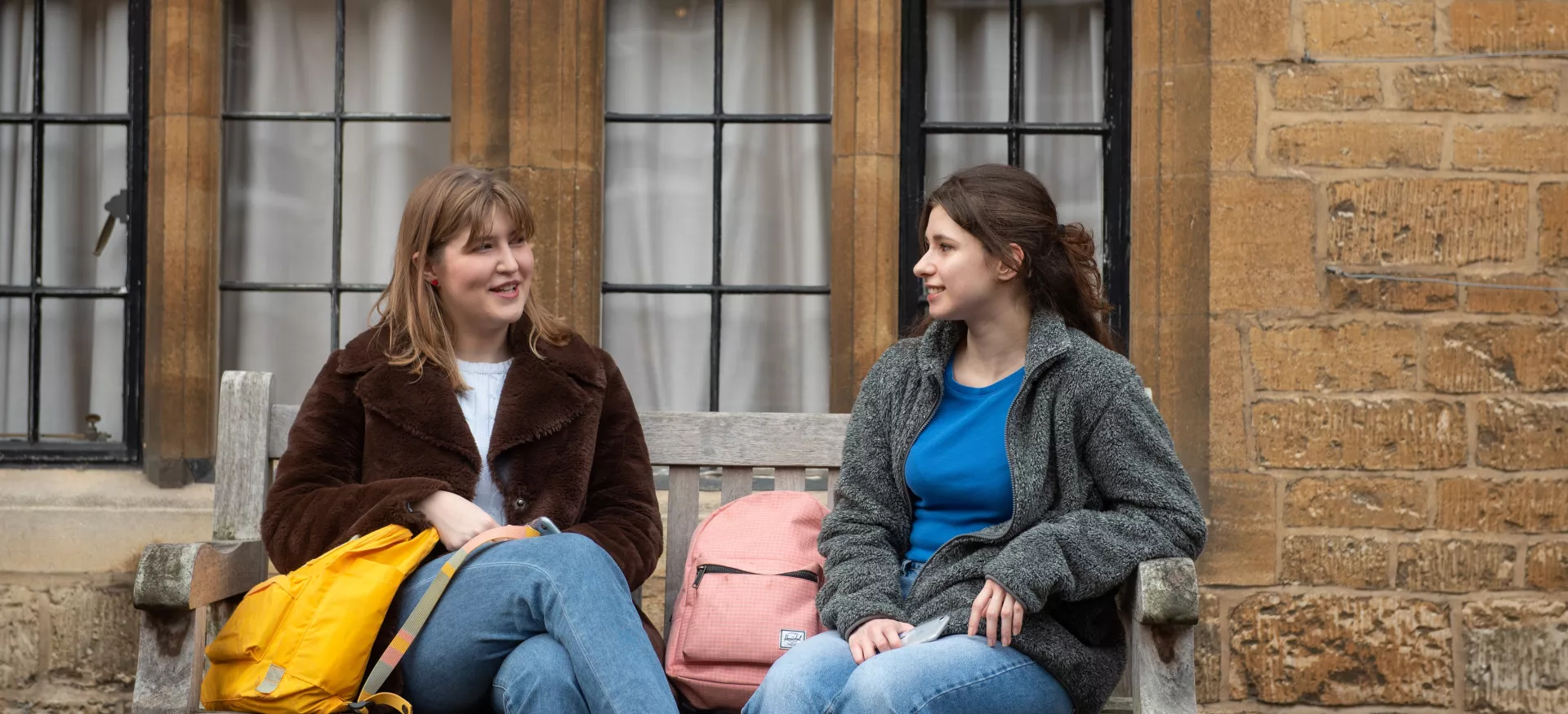 Two female students sit on a bench chatting in Trinity's library quad.