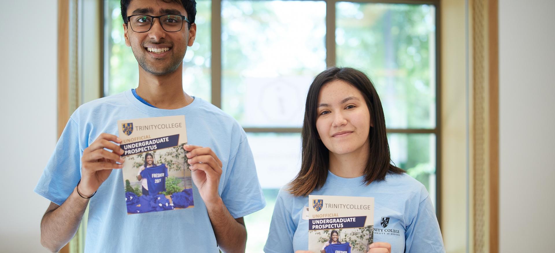 Two Trinity student ambassadors in matching blue shirts hold up undergraduate prospectuses.