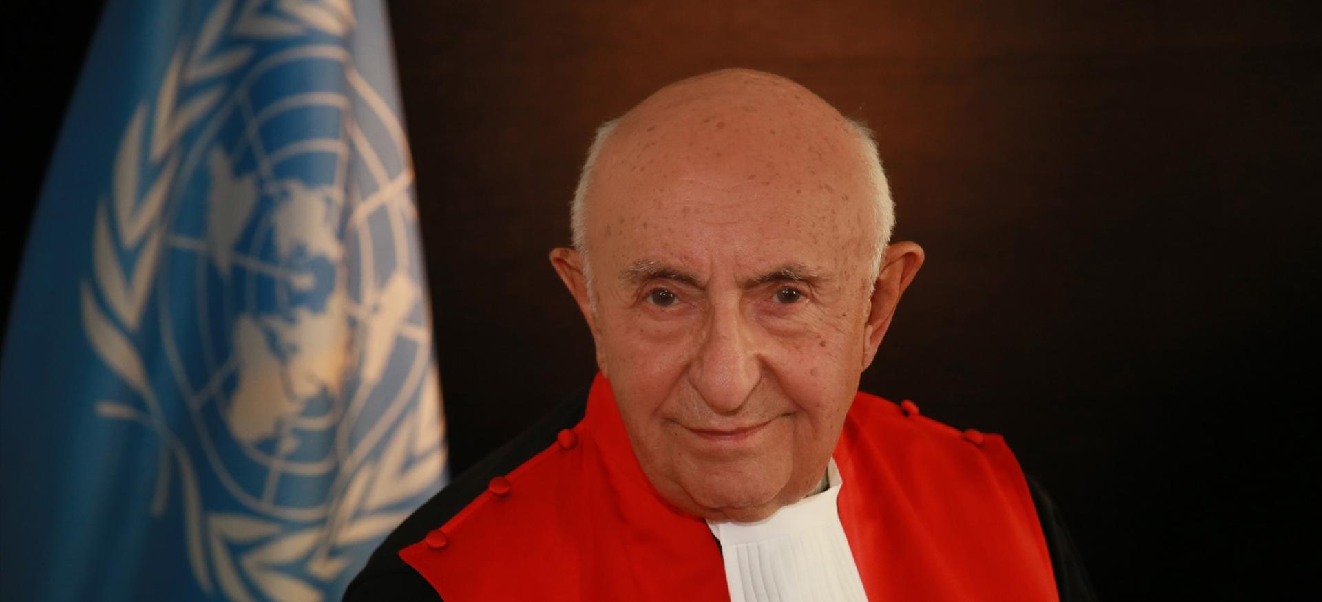 Judge Ted Meron wears his international criminal court robe and stands in front of a United Nations flag.