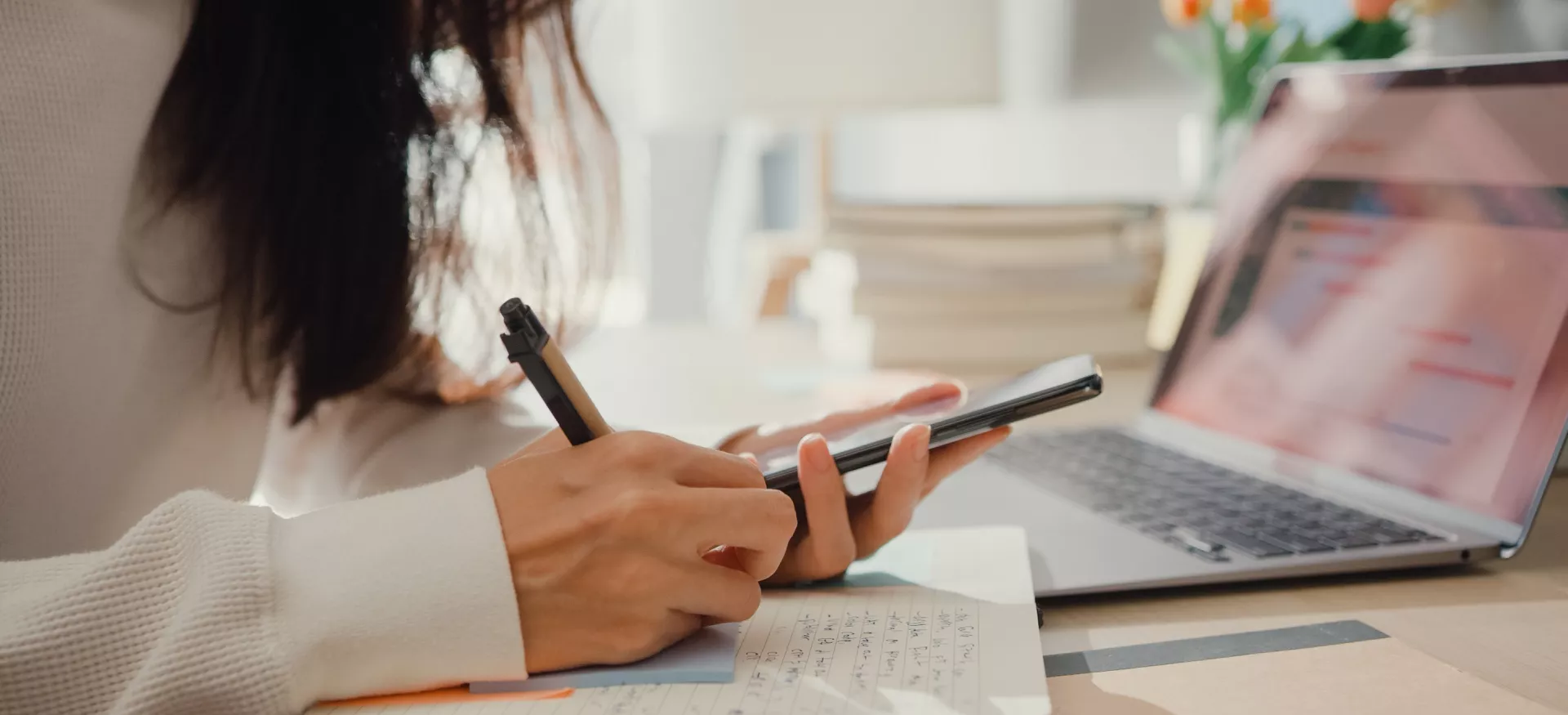 A female student sits at a computer taking notes and consulting her phone as she manages her budget.