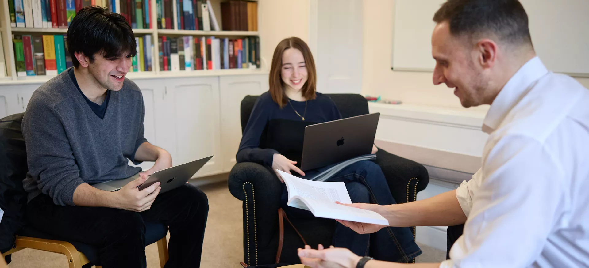 A male and a female student sit during a tutorial; their tutor gestures at a reading list while they look at their computers.