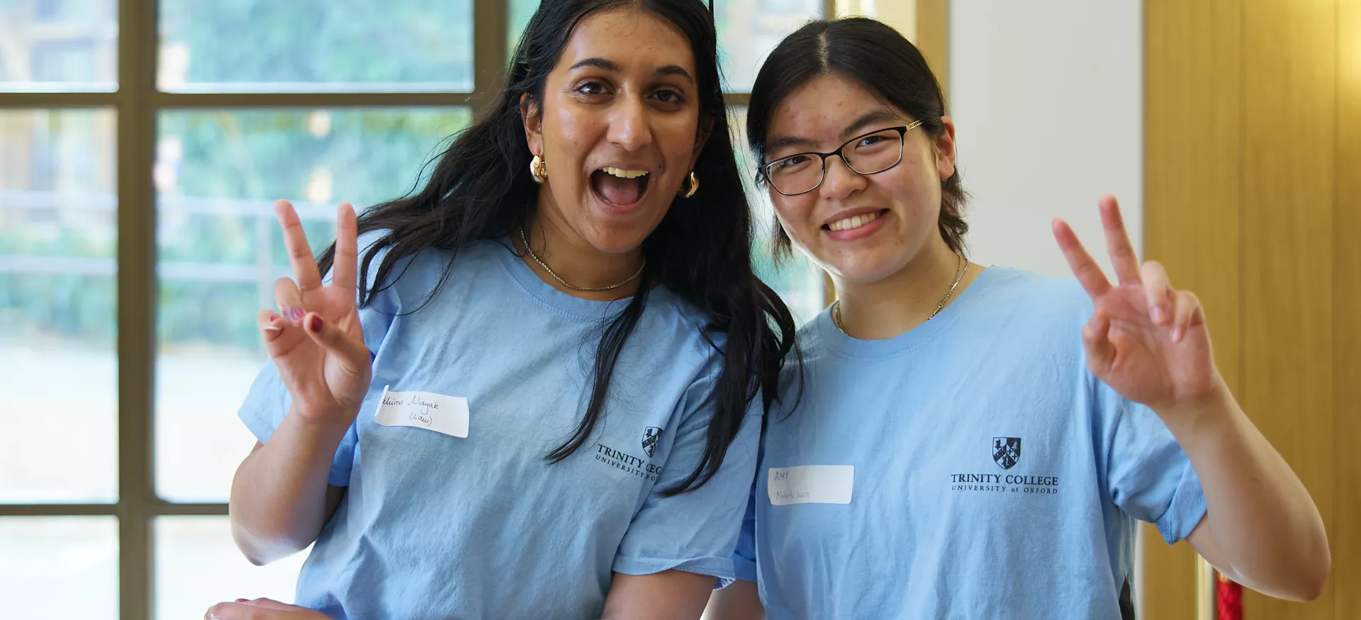 Two female students in matching light blue Trinity student ambassador t-shirts give peace signs in the cafe at open days.