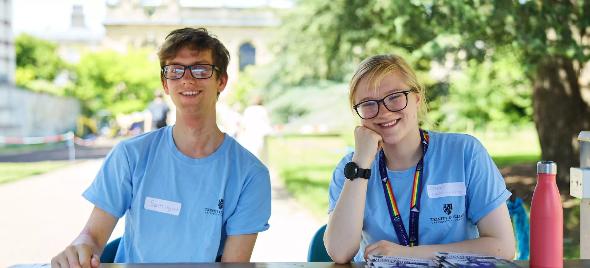 A male student and a female student sit in the front quad of Trinity welcoming visitors at an Open Day; they are wearing matching blue t-shirts.