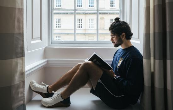 A male student reads on the window seat of his room with a view of Garden Quad out the window.