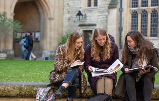 Three female Trinity college postgraduate students sit consulting books in one of the college quads.