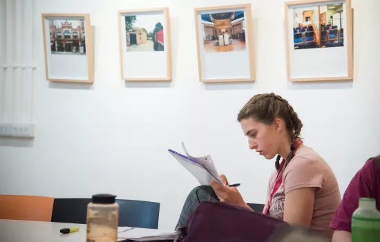 A female student writes in a notebook in a room with pictures hanging on the walls in the background.
