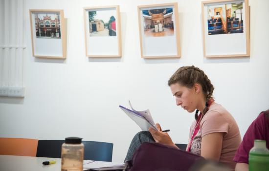 A female student writes in a notebook in a room with pictures hanging on the walls in the background.