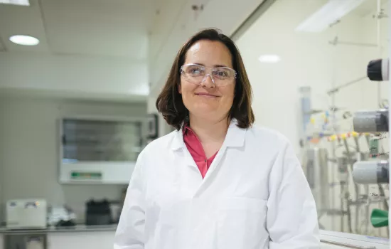 Charlotte Williams stands in her lab, wearing goggles and lab coat.