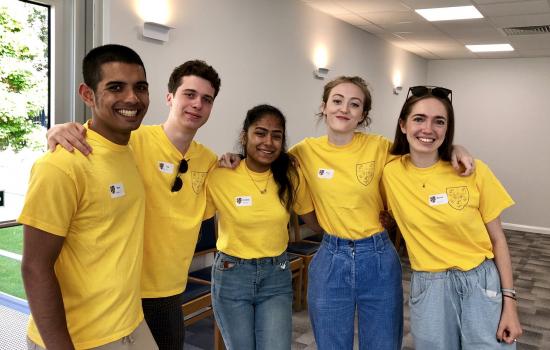 A group of five male and female Trinity students in yellow Open Days t-shirts stand with arms around each other at an Open Day.