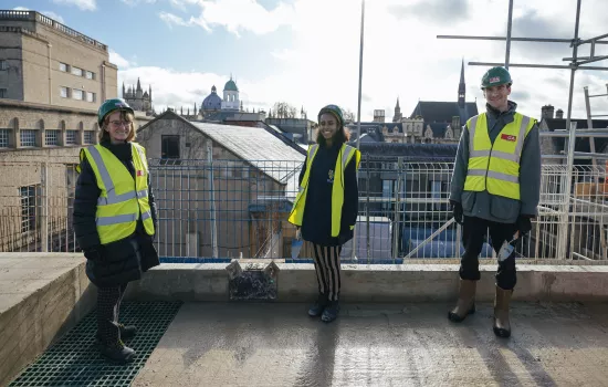 Trinity College's President, MCR President and JCR Presidents stand on the top of the Levine Building wearing hard hats and hi-vis jackets smiling. A partial view of the Oxford skyline is in the background.