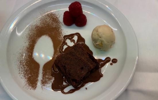 A plate with brownie drizzled with chocolate sauce and raspberries with a scoop of ice cream and sprinkled cocoa.