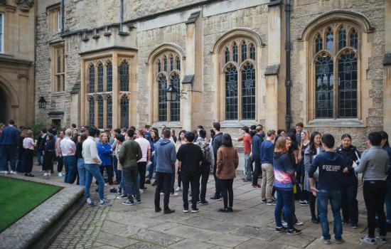 A large group of students stand talking outside the Trinity College dining hall.