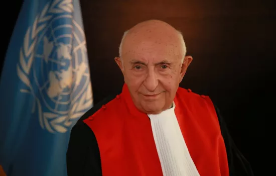 Judge Theodor Meron stands in his robes smiling in front of the flag of the United Nations.