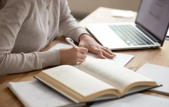 Shutterstock image of a person writing notes in a notebook with an open book and laptop next to them.