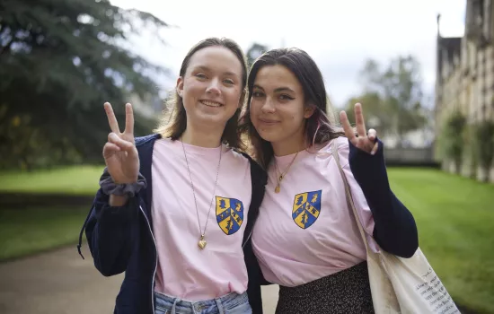 Two female Trinity students stand in the front quad of college making peace signs and smiling.