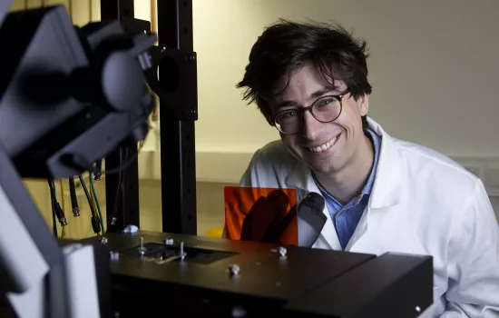 Trinity student Philippe Holzhey sits in front of a microscope; he is wearing a lab coat and goggles.