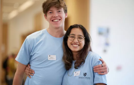A male and female Trinity college student stand in matching student ambassador t-shirts with their arms around each other smiling.