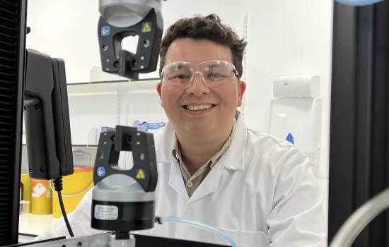 Trinity Dphil student Kam Poon sits at a lab microscope; he is wearing a white lab coat and goggles.