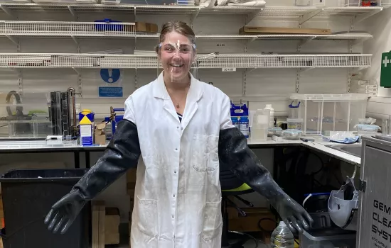 Trinity student Codrutza Dragu stands in the robotics lab wearing oversized lab coat and gloves, as well as safety goggles.