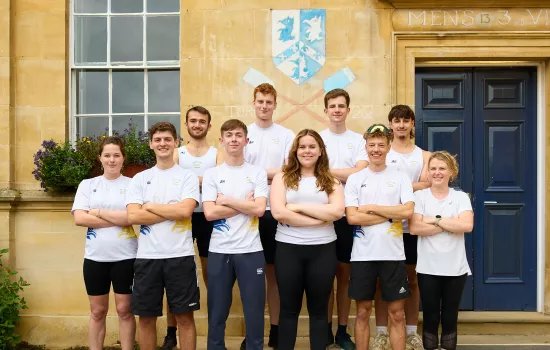 Nine members of the Trinity College Boat Club stand in matching kit in front of a building with rowing oars chalked on.