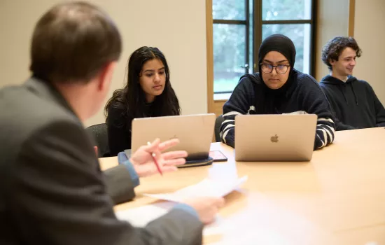 A group of students sit during a seminar at a desk with computers open.