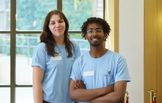 A male and female student in matching light blue student ambassador t-shirts stand in the cafe at open days.