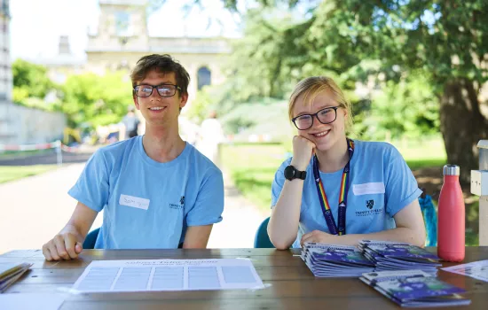 A male student and a female student sit in the front quad of Trinity welcoming visitors at an Open Day; they are wearing matching blue t-shirts.