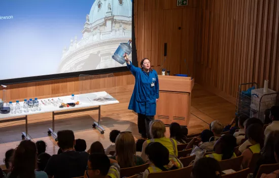Science presenter in a blue lab coat holding up a large water container during a live demonstration in Trinity College’s de Jager Auditorium. A table with experiment materials is set up beside her, and a projected image of Oxford’s Radcliffe Camera is displayed behind. Young students in high-vis jackets watch attentively from tiered seating.