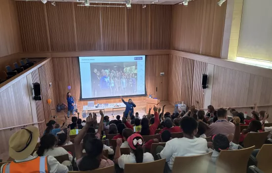 Large group of primary school children seated in Trinity's de Jager auditorium, many raising their hands during a lively science presentation. Two presenters in blue coats stand at the front near a projection screen displaying a photo of the Department of Chemistry team.
