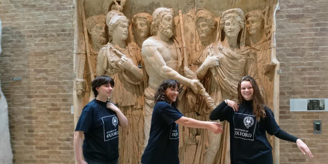 Three students wearing Oxford University T-shirts pose in front of a large sculptural wall. 
