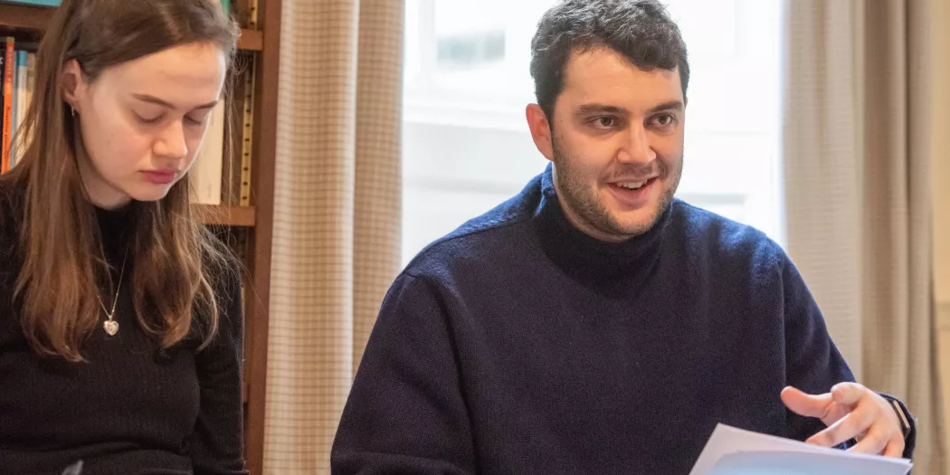 A male student smiles during a tutorial while a female student sits next to him consulting her papers. 