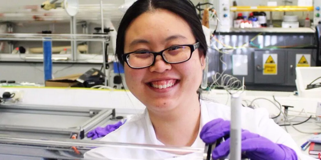 A female student smiles into the camera while holding some lab equipment; she is wearing a lab coat and purple safety gloves.