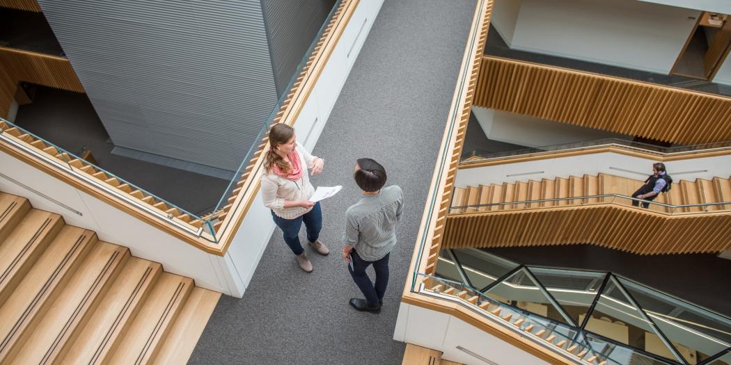 Two students stand talking on a staircase landing in the maths department. The image is taken from above.