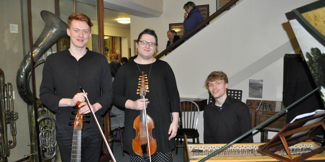 Two male students dressed in black hold violins while another student sits next to them at a piano in the Music Faculty at Oxford.