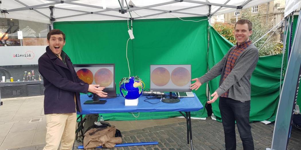 Two male students stand in front of a table in a marquee displaying their physics projects.