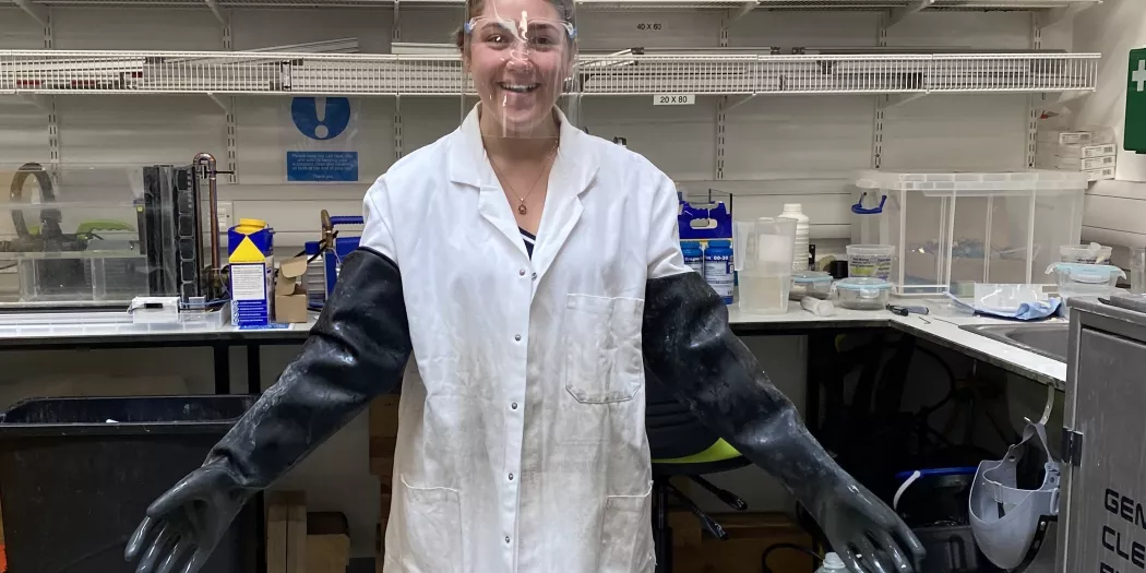 An engineering student stands in a lab coat and goggles with giant safety gloves on.