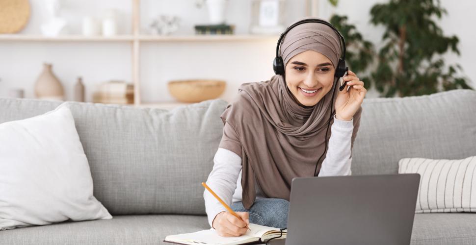 A female teacher wearing a head scarf and headset takes notes while looking at a computer during an online CPD session.