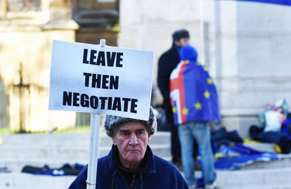 An older man in a coat and hat holds a sign saying 'Leave then negotiate;; in the background there is an EU flag.