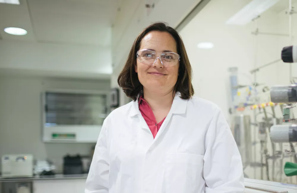 Charlotte Williams stands in her lab, wearing goggles and lab coat.